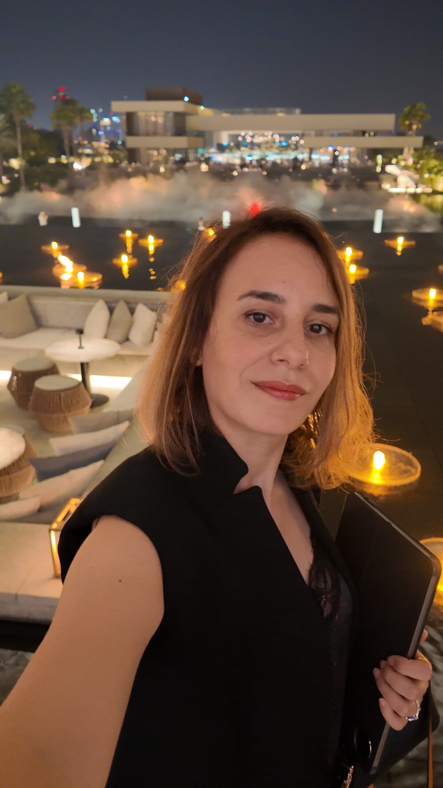 Sophia Benarafa holding a tablet on a Dubai waterfront terrace at night, with the Marina skyline behind her