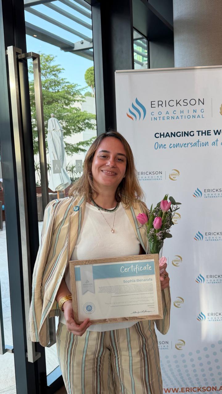 Sophia Benarafa holding her framed Erickson Coaching International certificate in front of the event banner